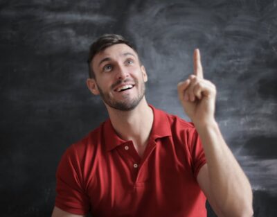Happy young man in a red polo shirt smiling and pointing upwards in front of a blackboard.