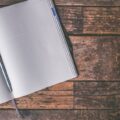 An open notebook with a blue pen lies on a rustic wooden table, inviting creativity and writing.
