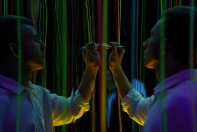 A man interacts with mirrored neon threads, creating vibrant reflections in a dark setting.