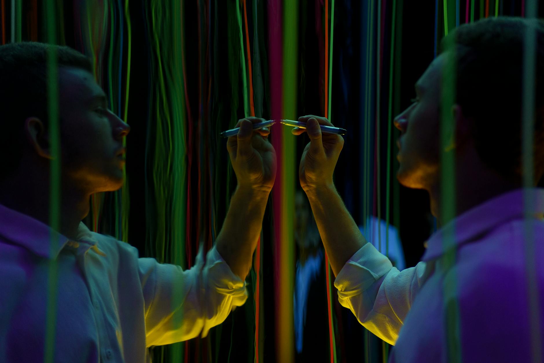 A man interacts with mirrored neon threads, creating vibrant reflections in a dark setting.