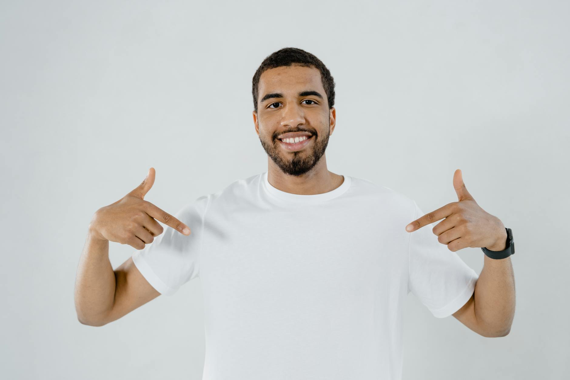 Portrait of a smiling man in a white shirt pointing at himself on a plain background.